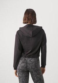 Hoodie nero accorciato con cappuccio, caratterizzato da una texture liscia. Abbinato a leggings scuri con motivo floreale, che mostrano un design aderente.
