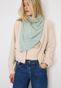 Donna bionda con giacca beige chiaro, sciarpa in maglia verde pallido, top beige e jeans blu, con una mano in tasca, che guarda verso il basso di lato.