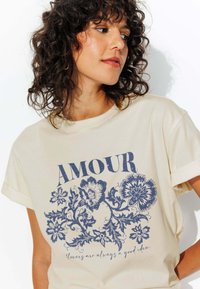T-shirt crème avec des manches roulées, présentant un graphique floral bleu et le texte "AMOUR". Tissu en coton doux, coupe décontractée.