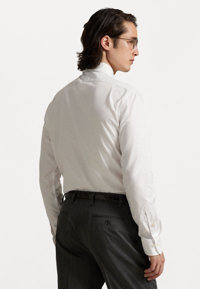 Valge nööpidega särk klassikalise kraega, millele on lisatud hallid ametirouhelised püksid, mis on siledast materjalist ja millel on küljetasku detail.