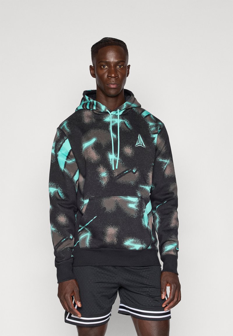 Sudadera con capucha en negro con patrones abstractos en turquesa y marrón, que cuenta con un bolsillo tipo canguro y cordones ajustables. Combinada con pantalones cortos negros.