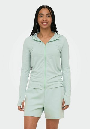 Hosszú fonott hajú nő világoszöld cipzáras kapucnis pulóvert és hozzá illő rövidnadrágot visel, áll és mosolyog egyszínű háttér előtt.