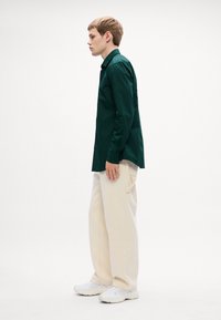 Donkergroene langemouwenshirt gecombineerd met lichtbeige wijde broek en witte sneakers, weergegeven op een effen witte achtergrond.