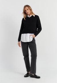 Zwarte gebreide trui met een V-hals, gelaagd over een witte blouse met kraag. Grijze rechte broek en zwarte instappers maken de outfit compleet.