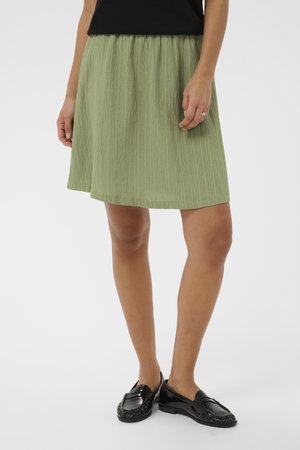 Kobieta w oliwkowej, teksturowanej spódnicy do kolan, czarnej bluzce i czarnych lordach, stojąca na jednolitym jasnym tle.