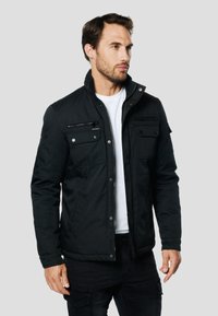 Melna jaka ar augstu apkakli, priekšā rāvējslēdzējs, divas krūšu kabatas un sānu kabatas. Raksturota ar gludu tekstūru un pogu akcentiem.