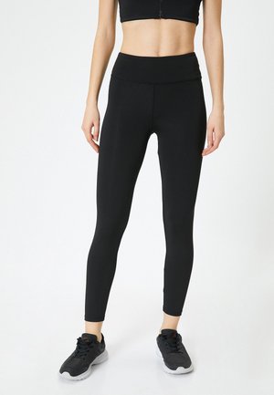 Leggings negros de cintura alta hechos de un tejido elástico. Diseño ajustado con una textura suave y costuras reforzadas. Llevados con zapatillas de deporte negras.