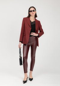 Rote, maßgeschneiderte Blazerjacke über einem schwarzen Spitzenoberteil, kombiniert mit hochglänzenden burgunderroten Leggings und schwarzen spitzen Pumps, während sie eine schwarze Handtasche hält.