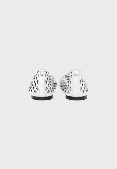Paire de chaussures blanches plates avec des découpes en forme de losange, semelles noires et petit logo métallique sur la languette du talon, vue de dos.