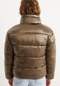 Person med skulderlangt hår iført en skinnende olivengrøn polstret jakke og lyseblå jeans, set fra ryggen mod en hvid baggrund.