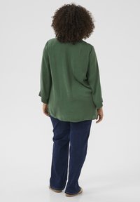 Blusa verde de manga larga con un corte relajado, tela texturizada y puños abotonados, combinada con jeans oscuros y zapatos de color claro.