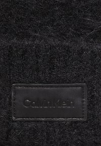 Tessuto nero texturizzato con un aspetto peloso presenta un'etichetta rettangolare in pelle nera con la scritta "Calvin Klein" in rilievo.