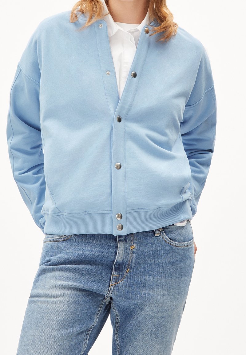 Personne portant un cardigan à boutons-pression bleu clair sur une chemise blanche et un jean bleu délavé, debout les mains dans les poches.
