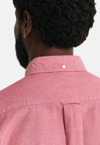 Roze overhemd met een knoop achterop en subtiele textuur. Bevat een plis voor extra bewegingsvrijheid en een enkele knoopdetail.