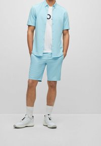 Hellblaues kurzärmeliges Hemd mit Knöpfen, weißes T-Shirt darunter mit schwarzem Logo, passende blaue Shorts, weiße Turnschuhe und Socken.