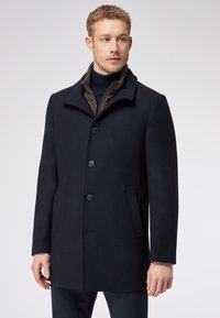 Cappotto in misto lana blu navy con un design strutturato, caratterizzato da un alto colletto, quattro bottoni, due tasche laterali e un dettaglio interno trapuntato.