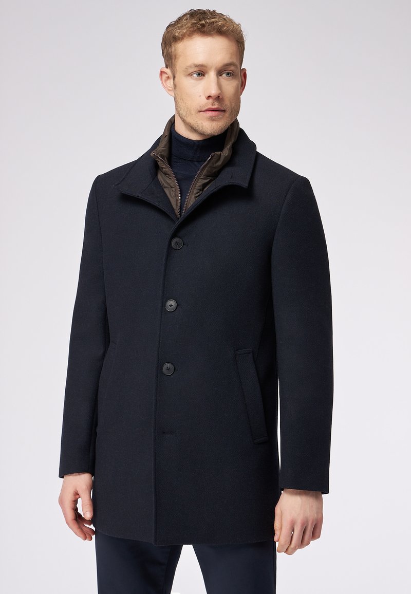 Cappotto in misto lana blu navy con un design strutturato, caratterizzato da un alto colletto, quattro bottoni, due tasche laterali e un dettaglio interno trapuntato.