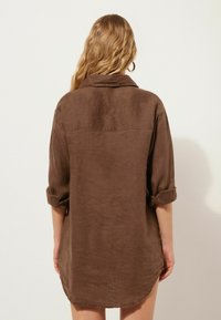 Braune Leinenbluse mit hochgekrempelten Ärmeln, einem Kragen und einem abgerundeten Saum. Der Stoff hat eine glatte Textur und einen dezenten Glanz.