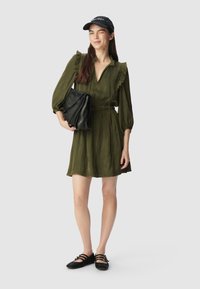 Zielono-oliwkowa sukienka z falbaniastym kołnierzykiem, elastycznym paskiem i długimi rękawami. Modelka trzyma czarną torbę z łańcuszkowymi akcentami, nosi czarną czapkę i płaskie buty.
