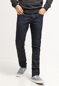 Jeans de mezclilla azul oscuro en corte slim, fabricados en algodón. Presentan un estilo estándar de cinco bolsillos y detalles de costura ligera. Combinados con zapatos grises tipo slip-on.