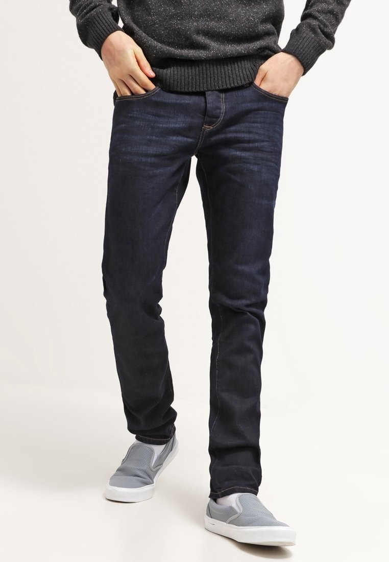 Jeans de mezclilla azul oscuro en corte slim, fabricados en algodón. Presentan un estilo estándar de cinco bolsillos y detalles de costura ligera. Combinados con zapatos grises tipo slip-on.