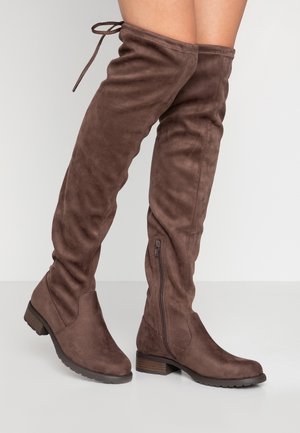 Bottes en daim marron au-dessus du genou avec talons empilés bas, fermetures éclair latérales et lacets à l’arrière sur les jambes d’une personne sur un fond uni.