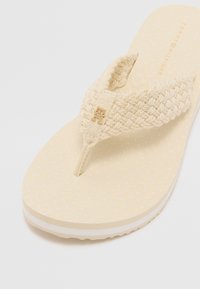 Beige flip-flop med et vævet rem, der har et subtilt geometrisk mønster på sålen og et lille guldlogo-accents på remmen.