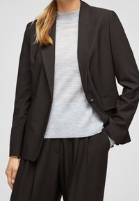 Blazer noir cintré avec un seul bouton, associé à un pull gris clair texturé. Col marqué et lignes épurées remarquables.