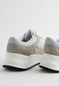 Helle graue Sportschuhe aus Mesh und Wildleder. Sie verfügen über eine robuste weiße Zwischensohle und eine strukturierte schwarze Außensohle. Gerundete Form, minimales Design.