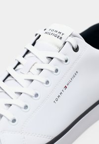 White leather sneaker with rounded toe, white laces, minimal branding on the sides, and a black rubber sole. Smooth texture with stitched detail.