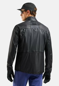 Mann trägt schwarze Mütze, Sonnenbrille, schwarze Handschuhe, leichte schwarze Jacke und blaue Hose, mit dem Rücken zur Kamera vor weißem Hintergrund.