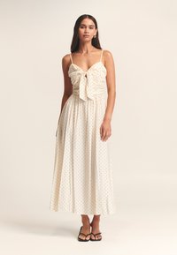 Woman wearing a cream-colored, sleeveless midi dress with black polka dots, a front tie at the bust, and black strappy sandals.