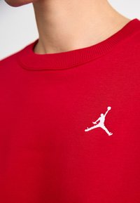 Rød hettegenser med ribbestrikket rund hals, med en hvit Jumpman-logo på forsiden. Stoffet ser glatt og lett ut.