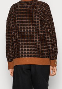 Maglione a maglia a righe nere e marroni con polsini e orlo a coste, indossato da una persona di spalle, abbinato a pantaloni neri.