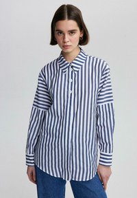 Camisa de botones a rayas que presenta franjas verticales alternas en azul marino y blanco, con cuello puntiagudo y mangas largas con dobladillo redondeado.