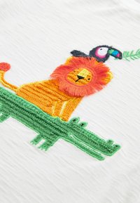 Broderet orange løve med en fluffy manke, der står på en grøn krokodille, med en farverig tukan, der holder et blad over dem.