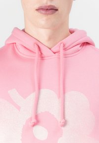 Rosa Kapuzenpullover aus weichem Stoff mit einem großen weißen Blumenmuster und passenden Kordeln. Das Design ist um den Halsausschnitt zentriert.