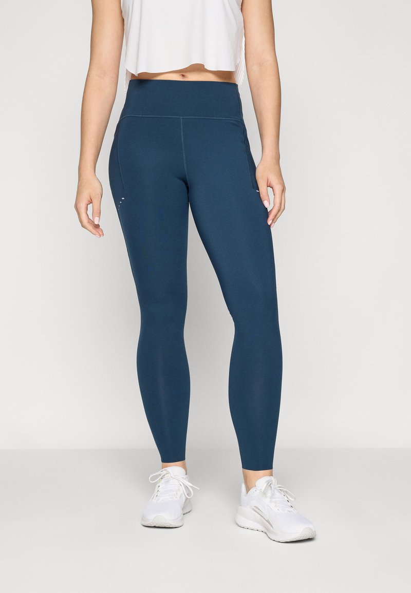Visoke sportske jeggings u tamnoplavoj tkanini, s glatkom teksturom i šivenim šavovima, u kombinaciji s bijelim sportskim cipelama.