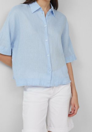 Vrouw draagt een loszittend lichtblauw overhemd met korte mouwen en witte opgerolde broek, staand tegen een effen achtergrond.