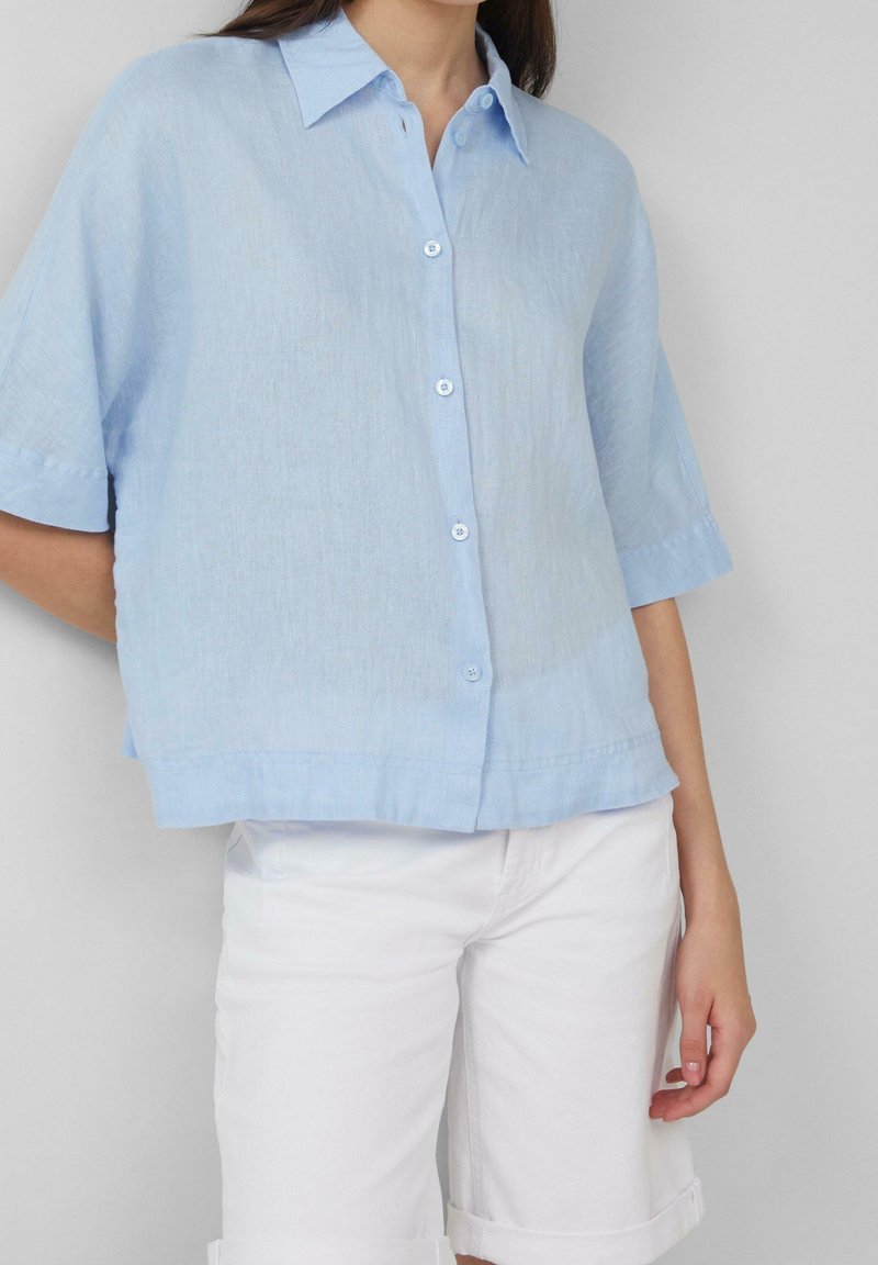 Femme portant une chemise ample bleu clair à manches courtes et un pantalon blanc retroussé, debout devant un fond uni.