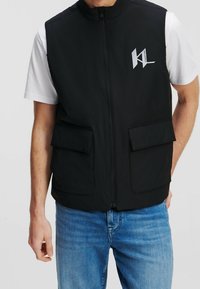 Svart ärmlös väst med framdragkedja, två fickor med lappar och logodetalj på vänster bröst. Bärs över en vit t-shirt och blå jeans.