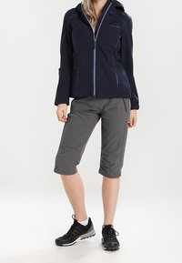 Marinblå softshell-jacka med dragkedja, grå cropped-byxor och svarta träningsskor. Outfiten har funktionell design och lätta material.