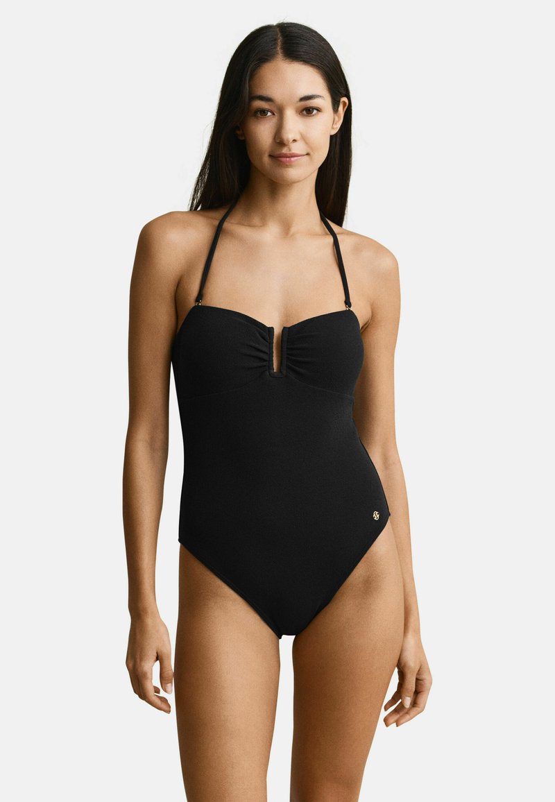 Femme aux longs cheveux noirs portant un maillot de bain une pièce noir avec des bretelles licou et une encolure en V au niveau de la poitrine.