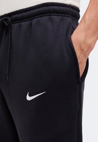 Κοντινό μαύρων παντελονιών φόρμας Nike με ελαστική μέση, κορδόνι, πλαϊνή τσέπη και λευκό λογότυπο Nike Swoosh με χέρι στην τσέπη.