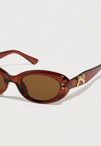 Γυαλιά ηλίου με καφέ χρωματισμούς, οβάλ φακούς, ευέλικτο πλαστικό πλαίσιο και χρυσή διακοσμητική φιόγκο στο σκελετό. Ημιδιαφανές σχέδιο από χελώνα.