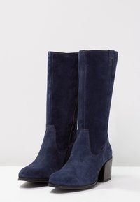 Ett par mid-kalv stövlar i marinblå mocka med blockklackar och sidodragkedjor på en vit yta.