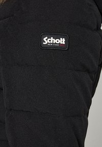 Veste matelassée noire avec un patch logo rectangulaire affichant "Schott New York NYC" en blanc et rouge. Tissu doux et texturé avec des coutures renforcées.