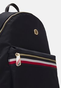 Granatowy plecak z nylonu o okrągłym kształcie, złotymi suwakami, przednia kieszeń z paskiem w paski w kolorze czerwonym, białym i granatowym oraz złotym emblematem logo.