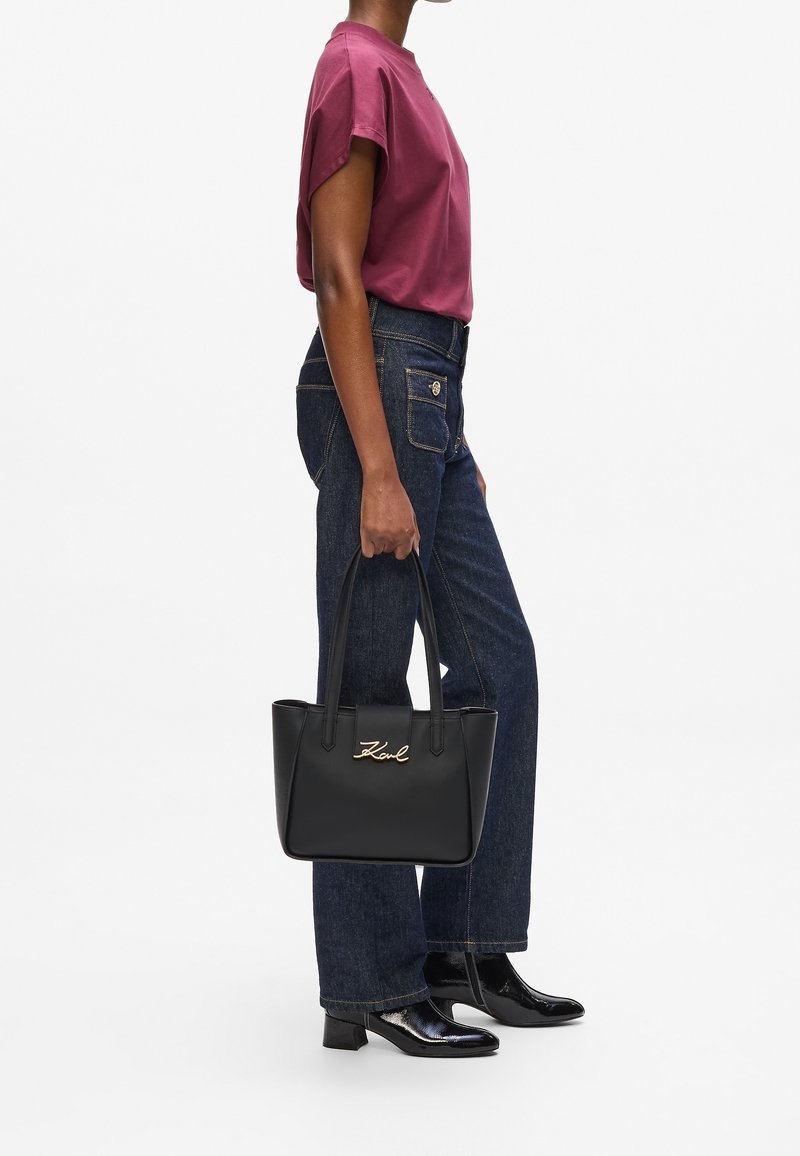 Femme en jean foncé et haut bordeaux tenant un sac à main en cuir noir avec le logo doré « Karl », portant des bottines noires à talons.