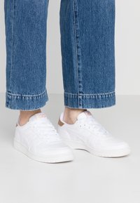 Weiße Sportschuhe mit einer strukturierten Oberseite, die einen hellbraunen Akzent an der Ferse, eine strukturierte Sohle und Schnürsenkel für einen sicheren Sitz aufweisen.